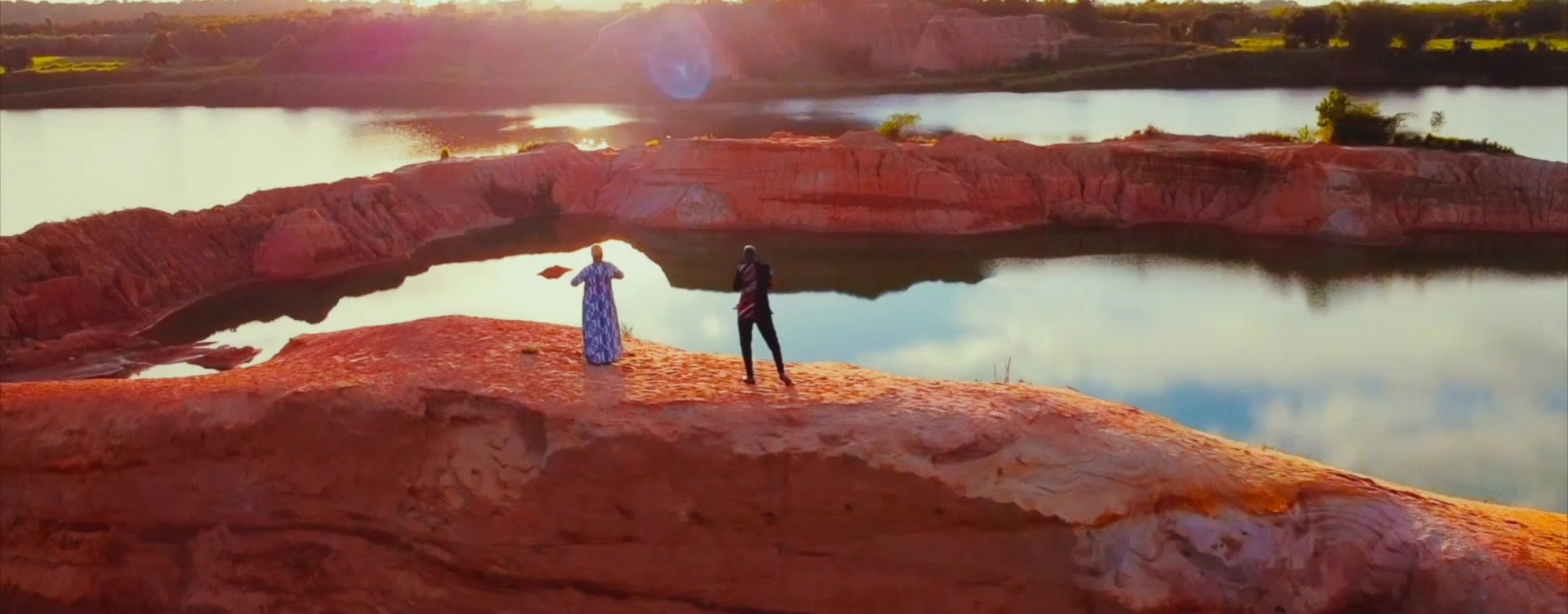 Two figures on a red rock arch above water at sunset